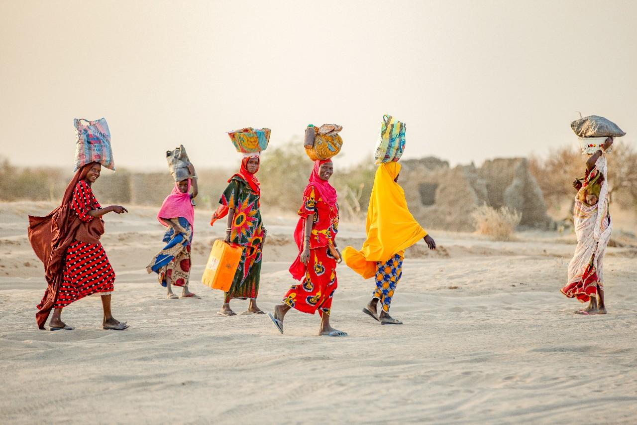 Women in brightly coloured clothing walk across sand carrying water vessels on their heads,
